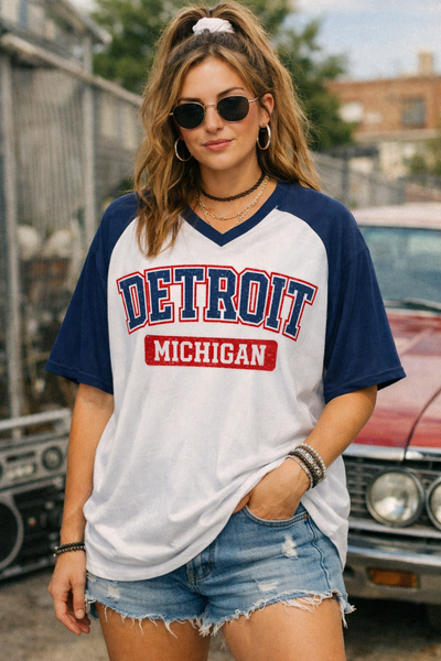 Woman wearing a 'Detroit Michigan' t-shirt with sunglasses and denim shorts, standing in front of a vintage car.