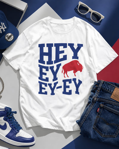 White t-shirt with 'HEY EY EY-EY' text and red graphic, surrounded by blue and white sneakers, sunglasses, and jeans on a colorful background.