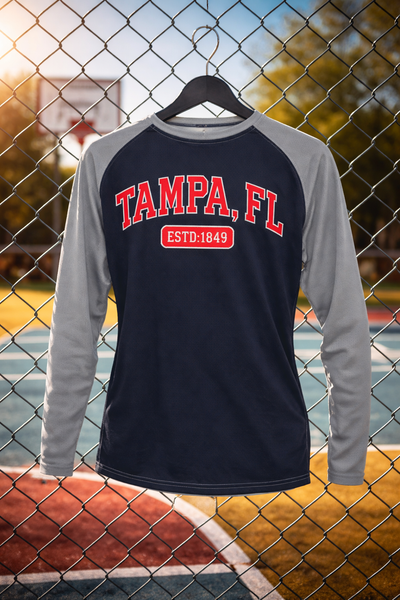 Long-sleeve shirt with 'Tampa, FL' text on a hanger against a chain-link fence background.