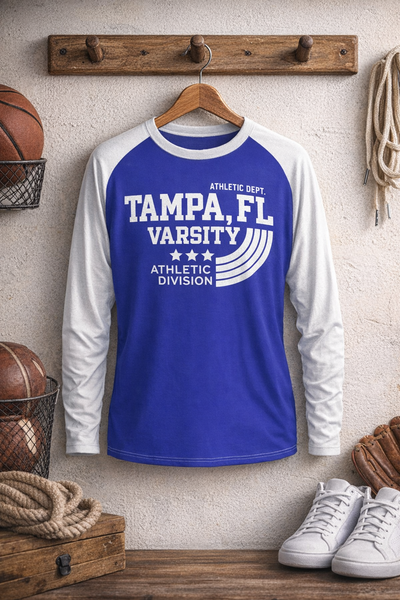 Blue and white long-sleeve shirt with 'Tampa, FL Varsity Athletic Division' text hanging on a wooden hook with sports equipment in the background.