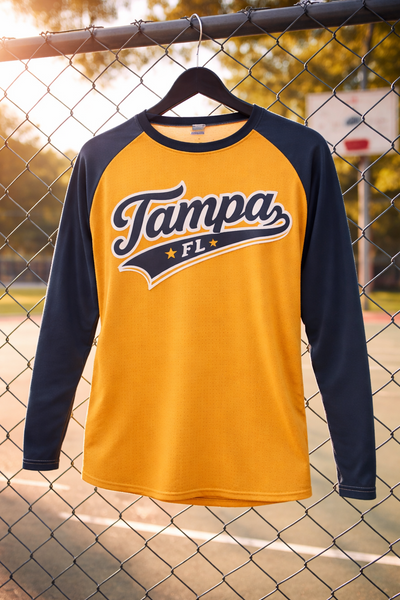 Yellow and navy long-sleeve shirt with 'Tampa, FL' text on a hanger against a chain-link fence background.