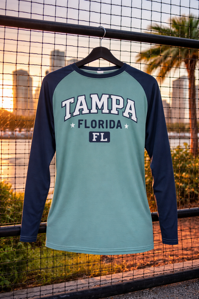 Teal and navy long-sleeve shirt with 'Tampa Florida FL' text on a hanger against a cityscape background.