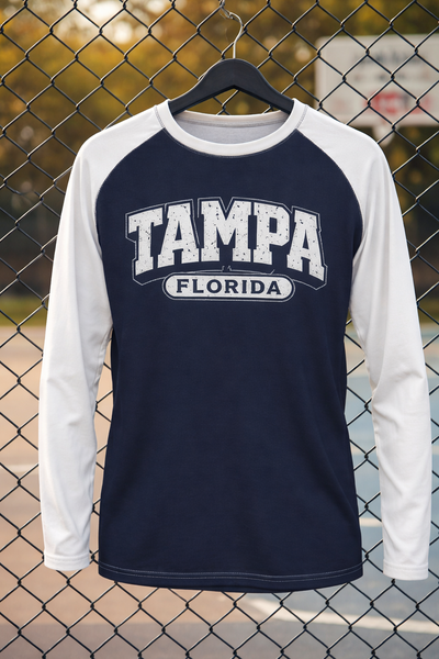 Navy and white long-sleeve shirt with 'Tampa Florida' text on a hanger against a chain-link fence background.