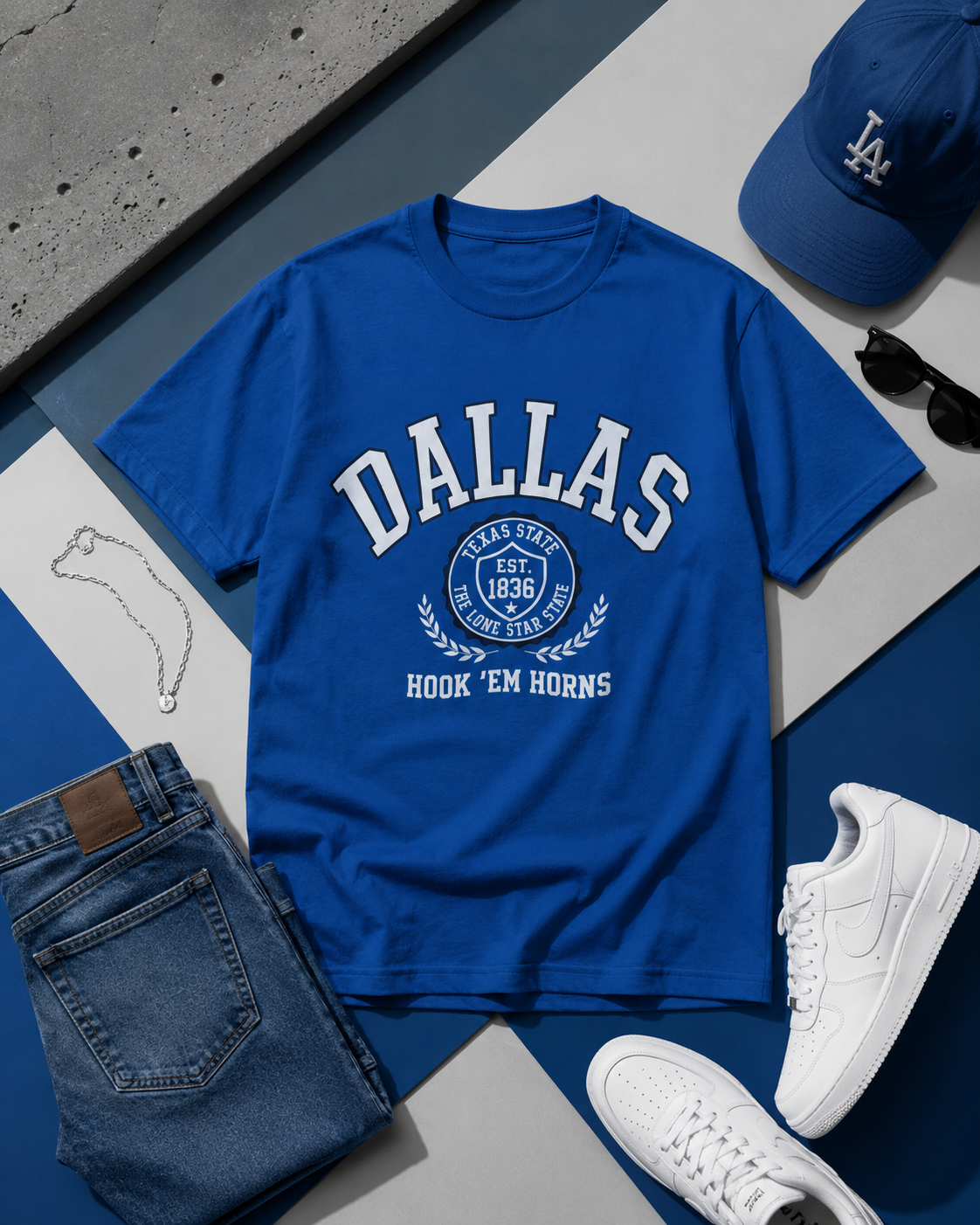 Blue 'Dallas' t-shirt with jeans, sneakers, and a cap on a blue and gray surface