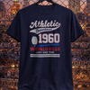 Navy blue t-shirt with 'Athletic Department 1960 Buffalo Bills Just One Time' text and graphic on a hanger against a cityscape background.