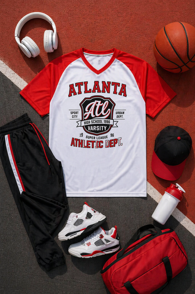 Red and white sports-themed t-shirt with athletic branding, surrounded by black shorts, red and white sneakers, a red duffel bag, basketball, headphones, and a cap on a brown surface.