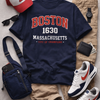 Navy t-shirt with 'Boston 1630 Massachusetts City of Champions' text, white and blue sneakers, red and white cap, beige pants, and accessories on a gray surface.