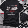 Black t-shirt with 'Boston Champs' text, surrounded by casual clothing and accessories on a gray surface.