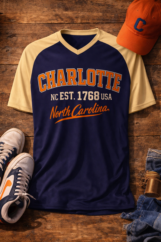 Navy and beige t-shirt with 'Charlotte, NC Est. 1768 USA North Carolina' text on a wooden surface with sneakers and a cap.