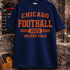 Navy blue t-shirt with orange text 'Chicago Football 1920 Soldier Field' on a hanger against a rustic background.