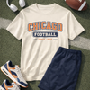 White t-shirt with 'Chicago Football' design, navy shorts, white sneakers, headphones, and a football on a green surface.