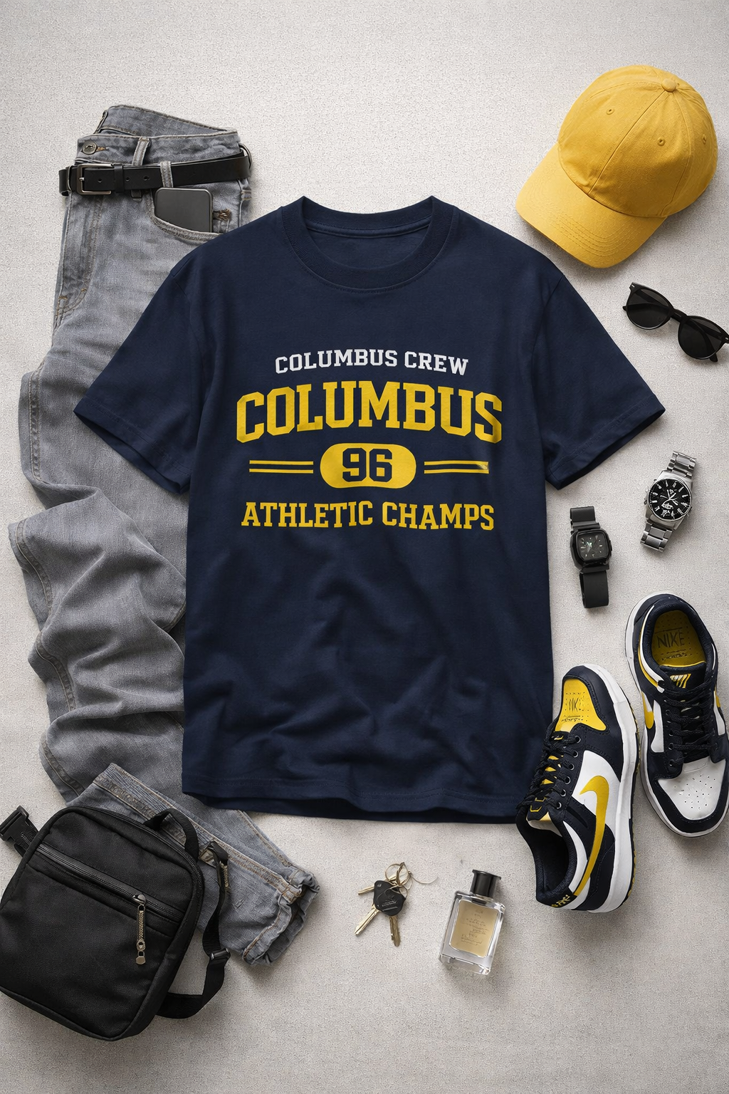 Navy t-shirt with 'Columbus Crew Columbus 96 Athletic Champs' text, surrounded by casual clothing and accessories on a light gray surface.