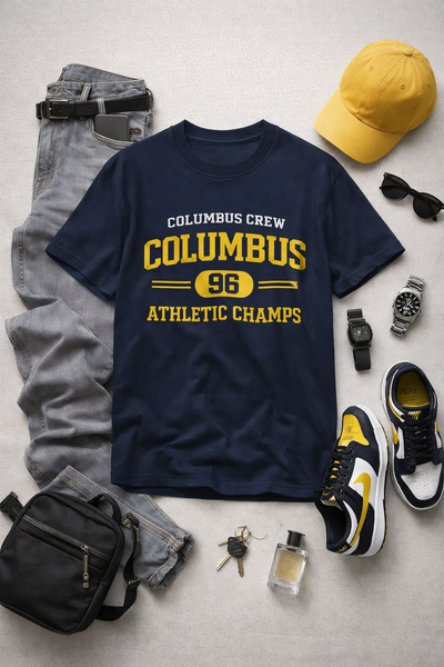 Navy t-shirt with 'Columbus Crew Columbus 96 Athletic Champs' text, surrounded by casual clothing and accessories on a light gray surface.