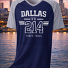 Navy and gray t-shirt with 'Dallas TX 214 North Texas' text on a hanger against a cityscape background.