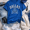Collection of clothing and accessories including a blue 'Dallas' t-shirt, white sneakers, jeans, and a cap on a light gray background.