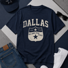 Navy blue t-shirt with 'Dallas' and star design, paired with jeans, white sneakers, and a cap on a gray surface.
