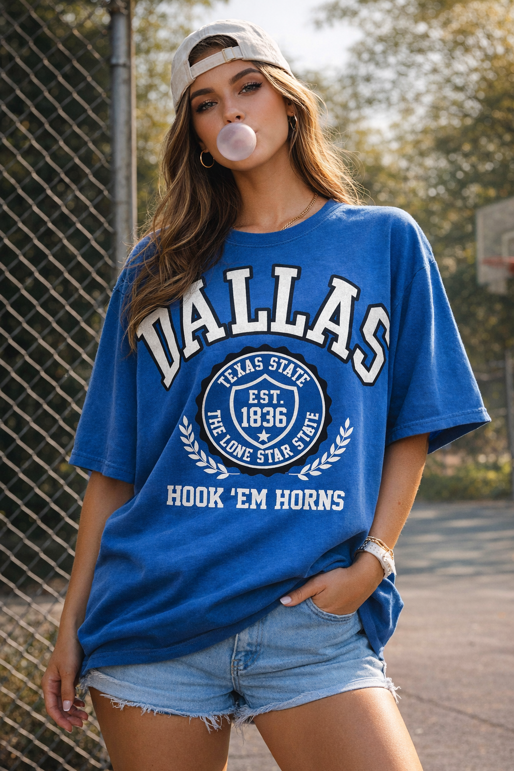 Woman wearing a blue 'DALLAS' t-shirt blowing bubble gum outdoors.