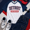 Detroit-themed clothing and accessories including a t-shirt, sneakers, shorts, and a cap on a red, white, and blue striped background.