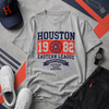 Gray t-shirt with 'Houston' text and sports-themed design, surrounded by a cap, sneakers, and watch on a patterned surface.