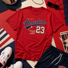 Red t-shirt with 'Houston 23' design, navy shorts, white sneakers, cap, and sunglasses on a colorful background.