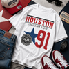 White t-shirt with 'Houston Lone Star State 91' design, surrounded by jeans, sneakers, cap, and sunglasses on a textured surface.