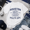 White t-shirt with 'Houston Athletic Dept.' design on a gray surface with jeans, sneakers, and a cap.