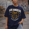 Person wearing a navy blue t-shirt with 'Los Angeles Dept. California' print, standing against a graffiti-covered wall.
