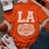 Orange t-shirt with 'Los Angeles' design, white sneakers, jeans, basketball, and cap on a textured surface.
