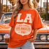 Woman wearing an orange 'Los Angeles' t-shirt in front of a classic car with palm trees in the background.