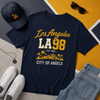 Navy t-shirt with 'Los Angeles LA98 City of Angels' design, surrounded by a cap, sunglasses, sneakers, and jeans on a multicolored background.