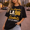 Woman wearing a black t-shirt with 'Los Angeles LA 98' text and palm tree design, standing on a street.