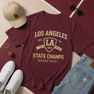 Maroon t-shirt with 'Los Angeles State Champs Basketball' text, beige cap, white sneakers, sunglasses, and keychain on a red surface.