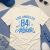 White t-shirt with 'Los Angeles 84 Athletic' text, blue cap, sunglasses, and keychain on a geometric patterned surface.