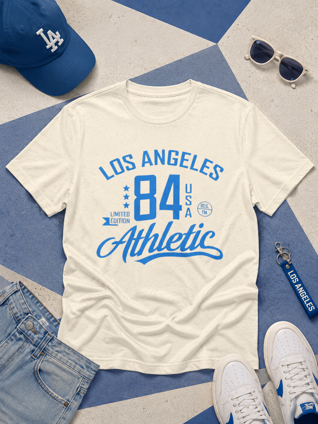 White t-shirt with 'Los Angeles 84 Athletic' text, blue cap, sunglasses, and keychain on a geometric patterned surface.