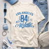 White t-shirt with 'Los Angeles 84 Athletic' design, blue cap, jeans, sneakers, and accessories on a light gray background.