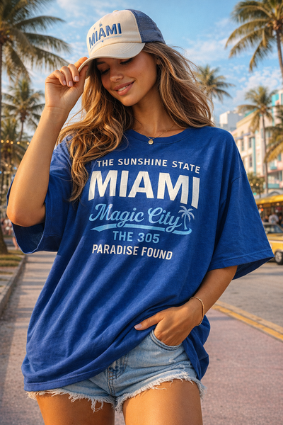 Woman wearing a blue 'Miami' t-shirt and cap on a sunny street with palm trees.