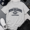 Gray t-shirt with 'New Orleans Louisiana' text, surrounded by casual items like a cap, sunglasses, and sneakers on a gray surface.