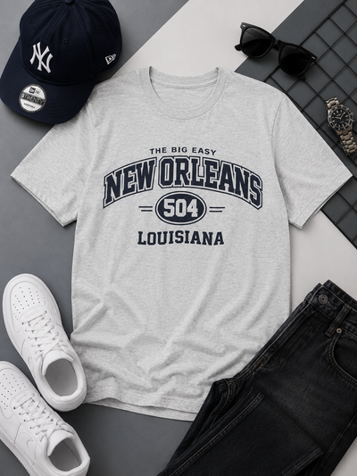 Gray t-shirt with 'New Orleans Louisiana' text, surrounded by casual items like a cap, sunglasses, and sneakers on a gray surface.