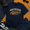 Navy t-shirt with 'New Orleans Athletics' text, black cap, white sneakers, sunglasses, and jeans on a two-tone background.
