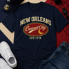 Navy t-shirt with 'New Orleans Crescent City' design, white sneakers, blue jeans, and a cap on a patterned background.
