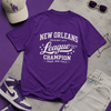 Purple t-shirt with 'New Orleans League Champion' text, surrounded by a cap, sunglasses, and sneakers on a gray and purple surface.