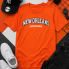 Orange t-shirt with 'New Orleans Louisiana' text, black cap, sunglasses, white sneakers, and black pants on a geometric patterned background.