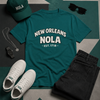 Teal t-shirt with 'New Orleans NOLA' text, green cap, white sneakers, black sunglasses, and black pants on a geometric patterned surface.