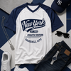 White and navy baseball-style t-shirt with 'New York' text, paired with white sneakers and a cap on a geometric patterned background.