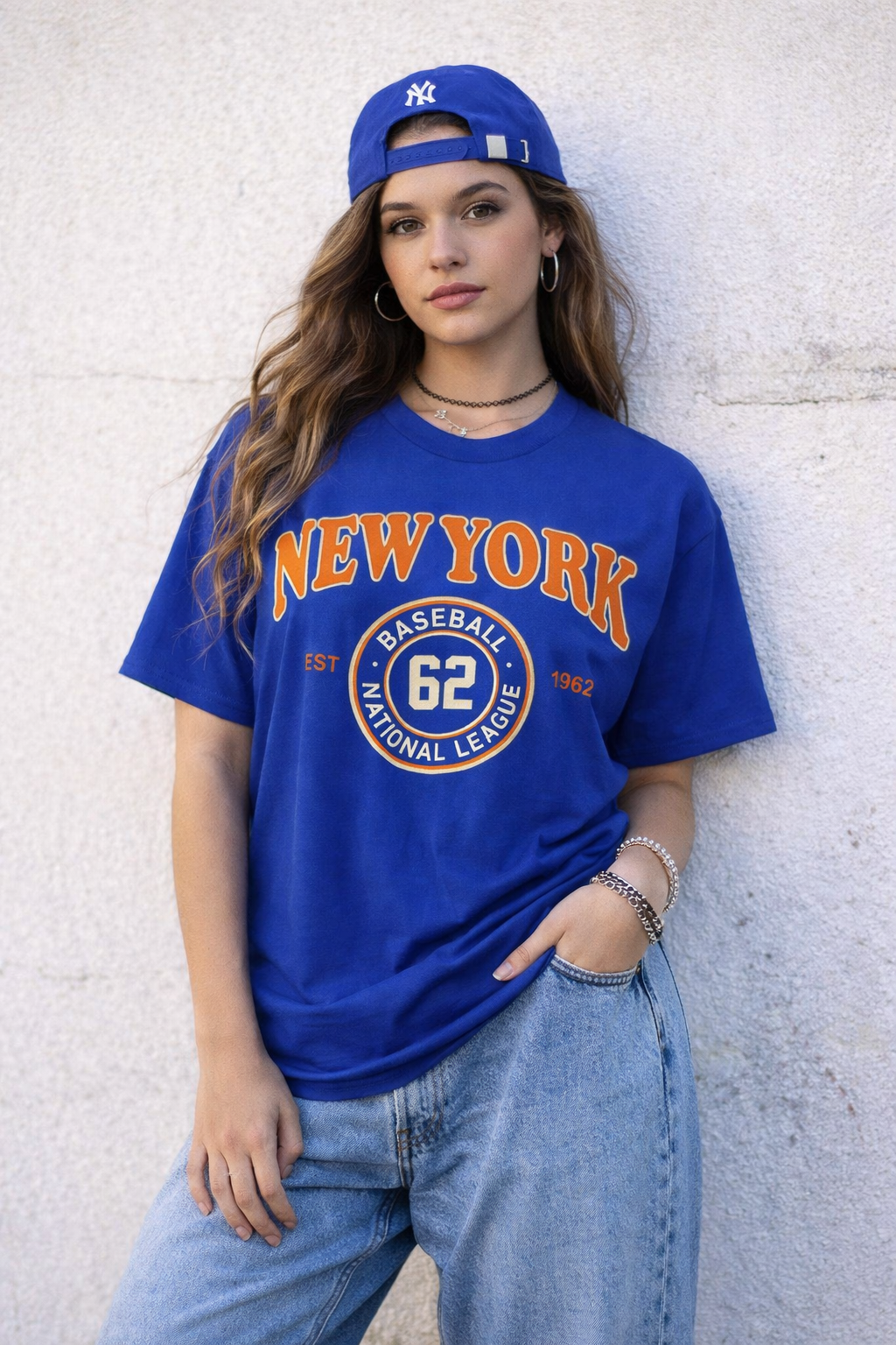 Person wearing a blue 'New York' t-shirt and cap against a white wall