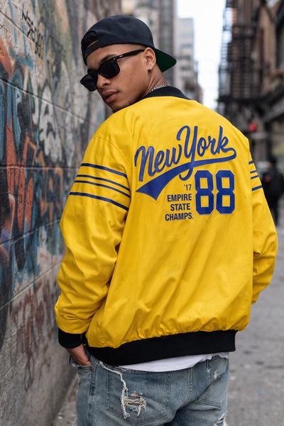 Person wearing a yellow 'New York' jacket with blue text, standing against a graffiti-covered wall.
