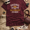 Maroon t-shirt with 'Original 1848 San Francisco' text, white sneakers, beige pants, and accessories on a wooden surface.