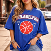 Woman wearing a blue 'Philadelphia' t-shirt with a basketball graphic on a city street.