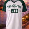 White and green baseball jersey with 'Philadelphia PA EST. 1933 Football' text on a hanger against a cityscape background.