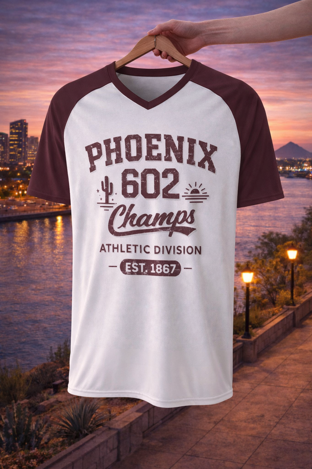 White and maroon baseball shirt with 'Phoenix 602 Champs' text on a hanger against a sunset cityscape background.